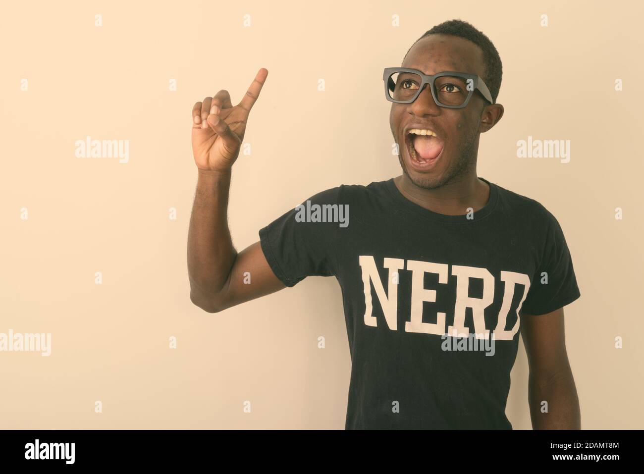 Foto de estudio de joven feliz negro africano geek hombre con Gran idea sonriendo y apuntando con el dedo hacia arriba mientras llevas Nerd camisa contra fondo blanco Foto de stock
