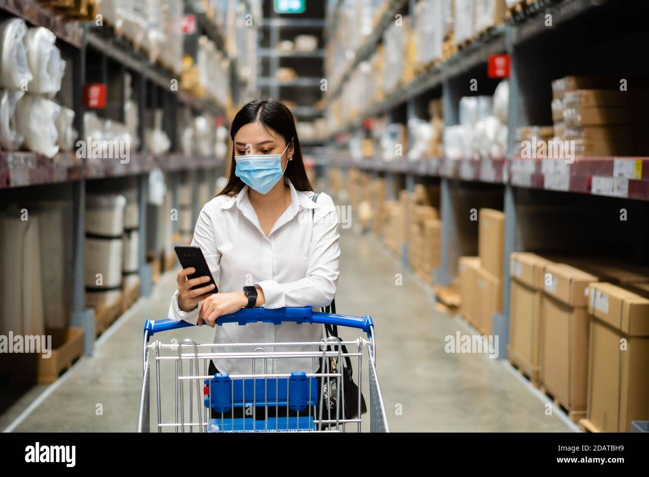 mujer con máscara médica mirando su teléfono móvil y. compras en el almacén durante la pandemia del coronavirus (covid-19) Foto de stock