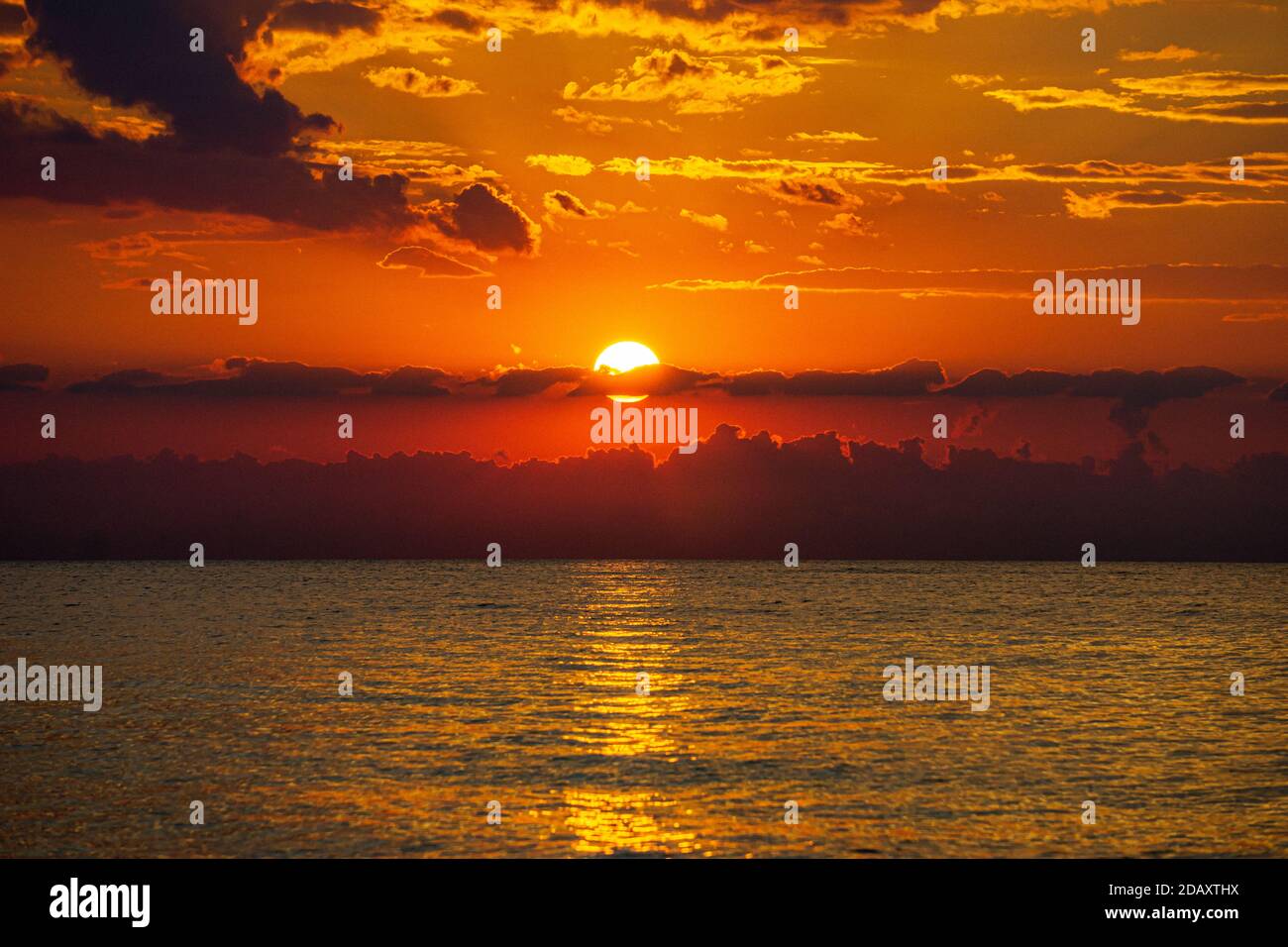 vista frontal de la puesta de sol sobre el mar con el reflejo de luz en el agua y en el fondo el sol y nubes al atardecer Foto de stock