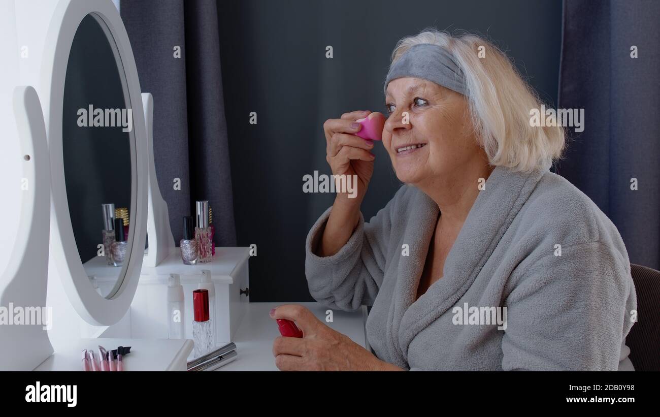 Abuela anciana cuidando de la piel, aplicando base antiarrugas con esponja,  poniendo maquillaje. Abuela de edad avanzada que hace procedimientos de  rehecho en el hogar. Cosméticos para jubilados Fotografía de stock -