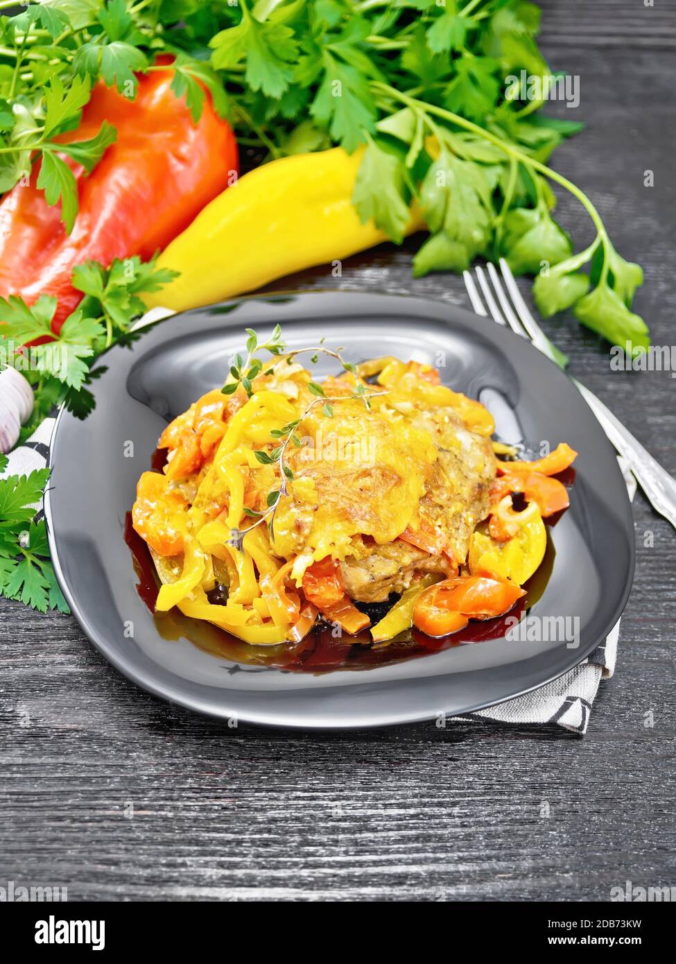 Pollo guisado con tomates, pimientos rojos y amarillos y queso en un plato sobre servilleta, tomillo, perejil y ajo sobre fondo de madera Foto de stock
