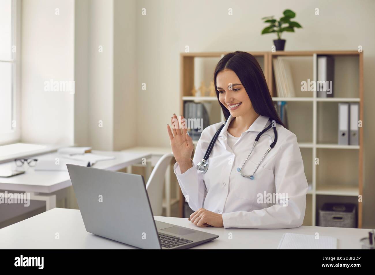 Médico amable en el consultorio del hospital haciendo videollamadas y agitando mano a la pantalla del portátil Foto de stock