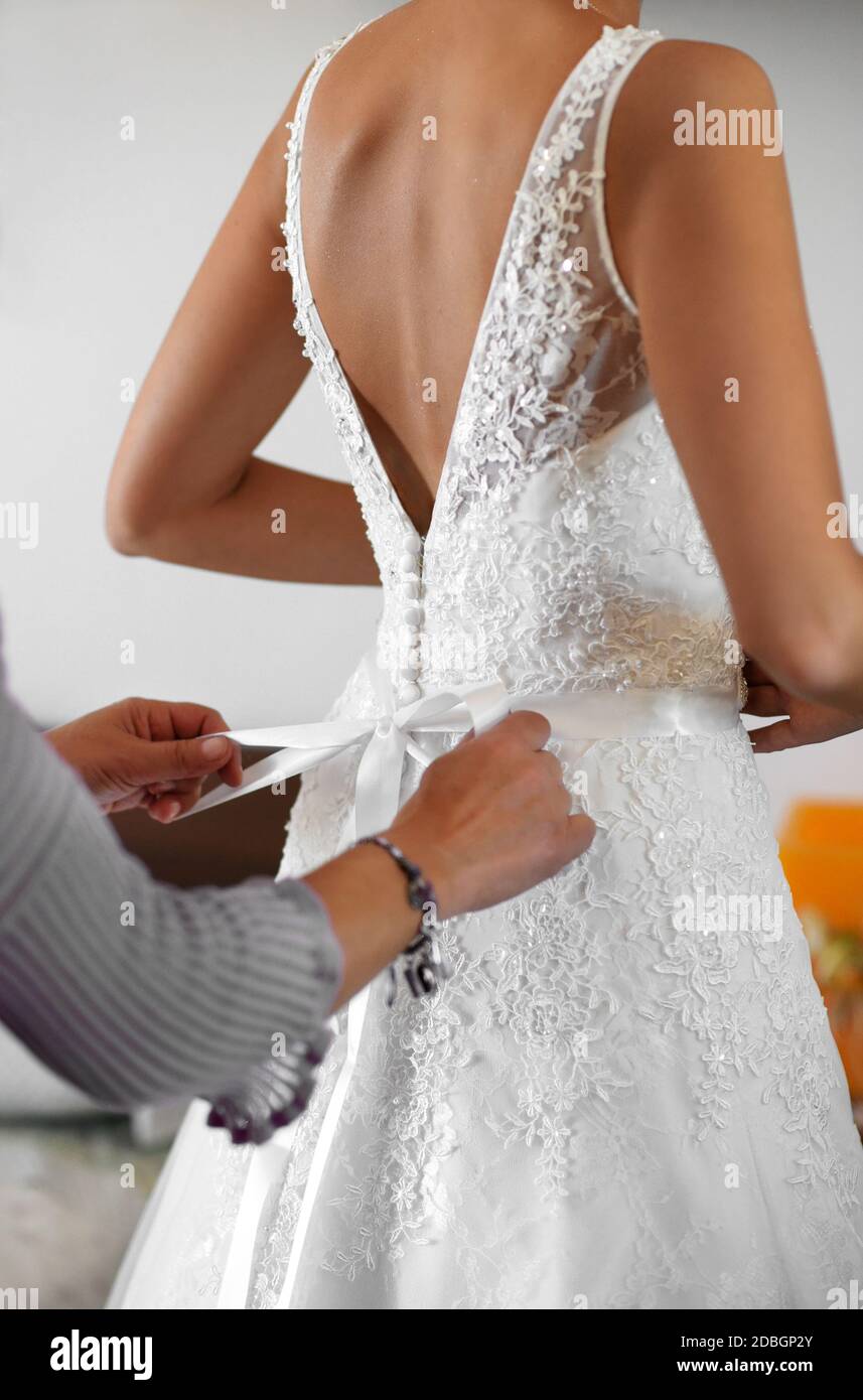 Criada de honor ayudando a la novia a vestirse en ella elegante bata blanca de boda que ata el arco en la parte de atrás de cerca en sus manos Foto de stock
