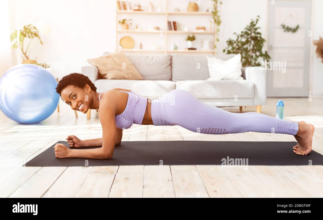 Deportes y pérdida de peso en casa. Mujer atlética afroamericana haciendo ejercicios en la estera de yoga en el interior de la sala de estar Foto de stock