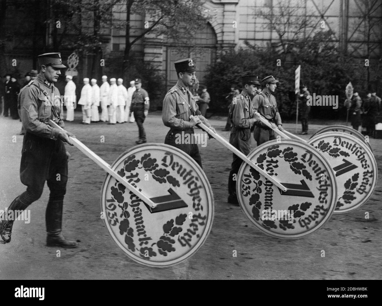 Cuatro miembros de SA durante una marcha promocional propagandística para la lotería de creación de empleo en Berlín. Hacen rodar grandes réplicas del Reichsmark a través de una plaza. Foto de stock