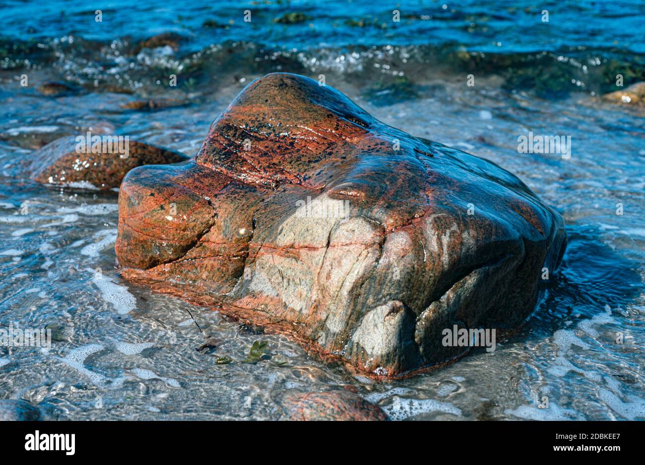 En la playa se encuentra una gran piedra rectangular de color marrón rojizo y es lavada por el agua de mar; se pueden ver pequeños whitecaps Foto de stock