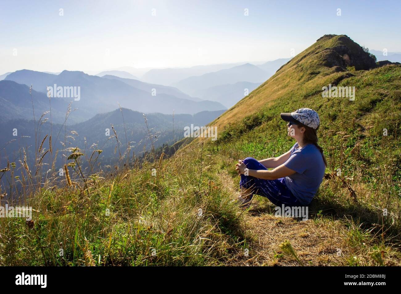 mujer joven en una gorra sentada entre las montañas y mirando el sol y la vista de la montaña. Paisaje, cresta de Aibga, caminata y trekking, viaje a Foto de stock