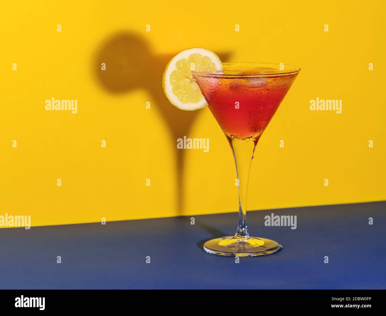 Cóctel rojo en un cristal que forma su sombra con un rodajas de limón sobre un fondo azul y amarillo Foto de stock