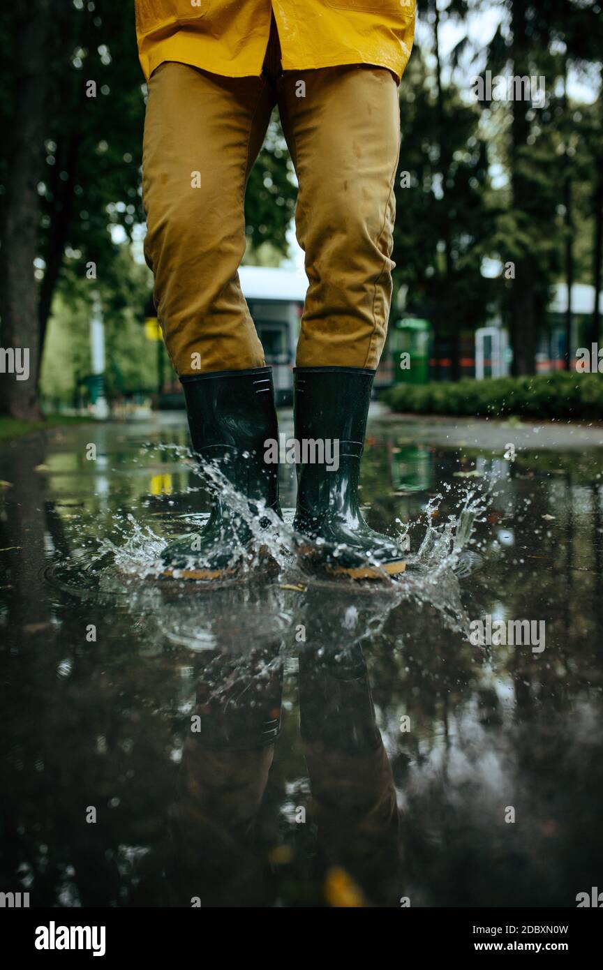 Hombre en la lluvia capa y botas de goma saltando en charcos, clima húmedo  en el callejón. El hombre posa en el parque de verano, día lluvioso.  Protección contra el agua, gotas