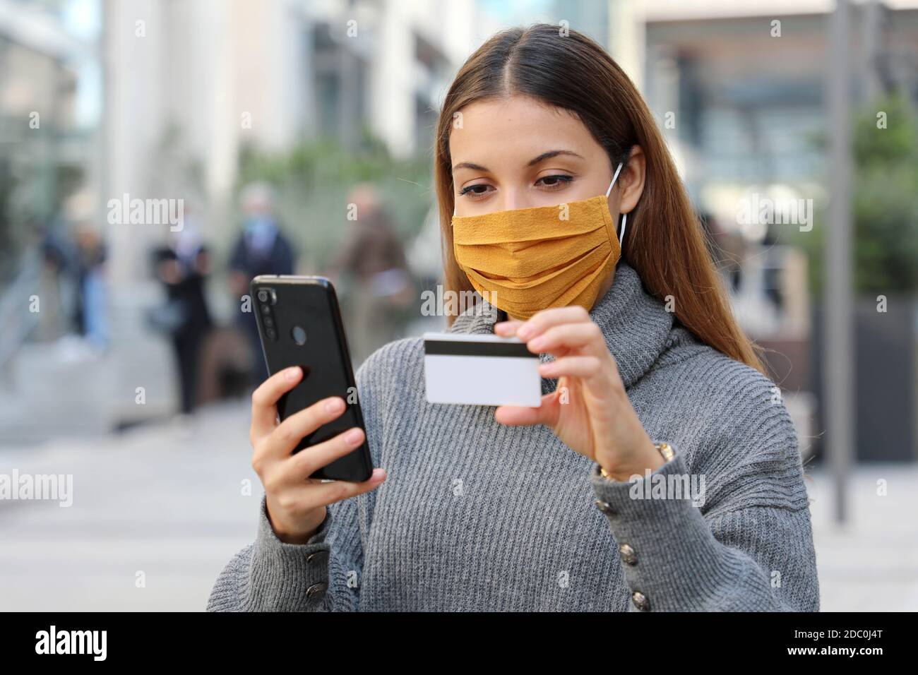 Retrato de joven hermosa mujer con máscara protectora chill out y realizar el pago a través de la aplicación de tarjeta de crédito y smartphone en la calle Foto de stock