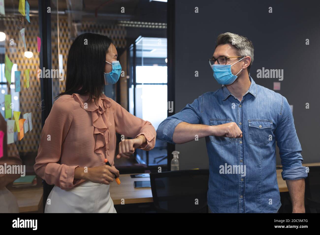 El hombre caucásico y la mujer asiática con máscaras faciales saludando a cada uno otros por tocar codos en moderno de Foto de stock