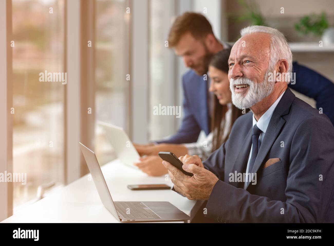 Gerente mayor usando un ordenador portátil con jóvenes coleguas en el fondo Foto de stock