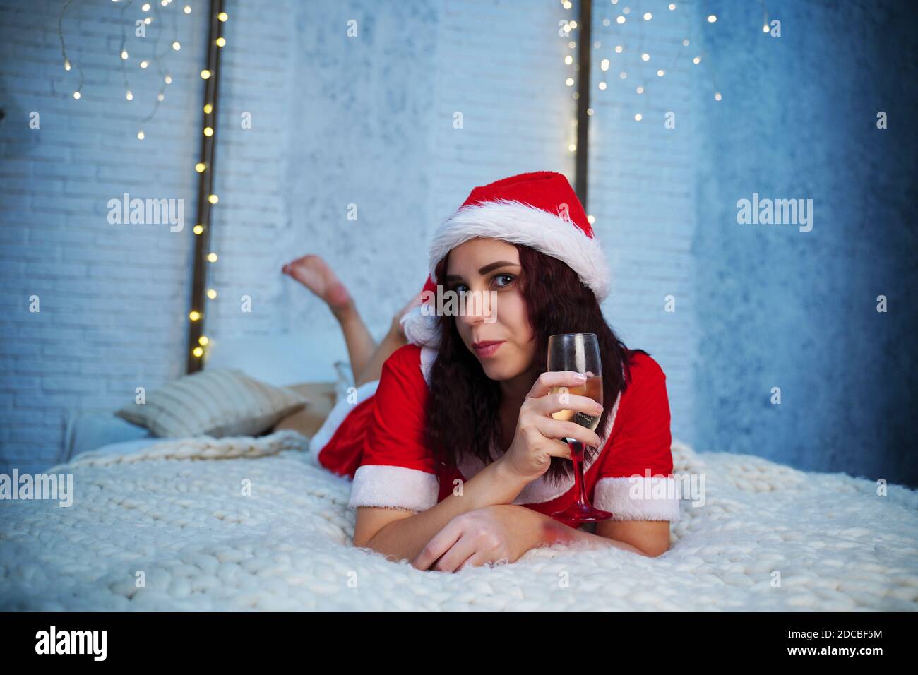 Mujer joven en traje de Santa Claus con una copa de champán tumbado en la  cama. Encantadora mujer descansando y celebrando la Navidad en casa.  Concepto de vacaciones Fotografía de stock -