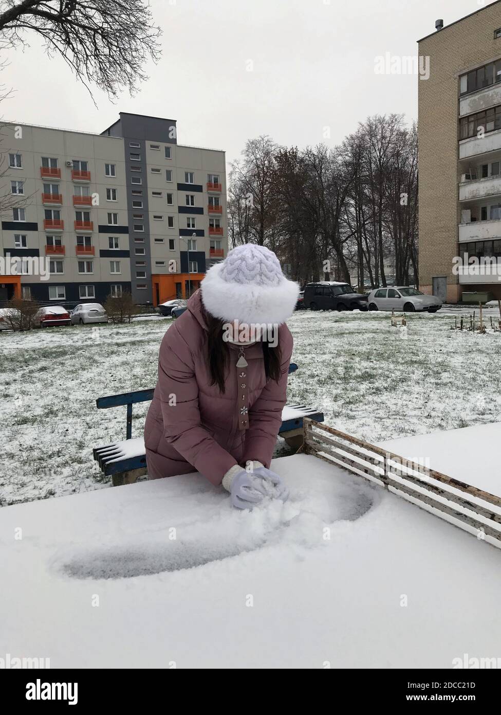 Chica con ropa de invierno para una caminata. Sostiene una bola de nieve en  sus manos. De pie cerca de una mesa de tenis de hormigón. Todo está  cubierto con la primera