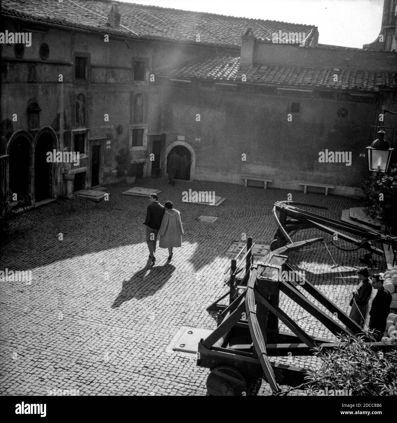 Un romántico paseo en pareja que se sostiene de la mano a través de una soleada plaza adoquinada en Roma, pasando por algunos arcos gigantes, mientras un rayo de sol se derrama. Foto de stock