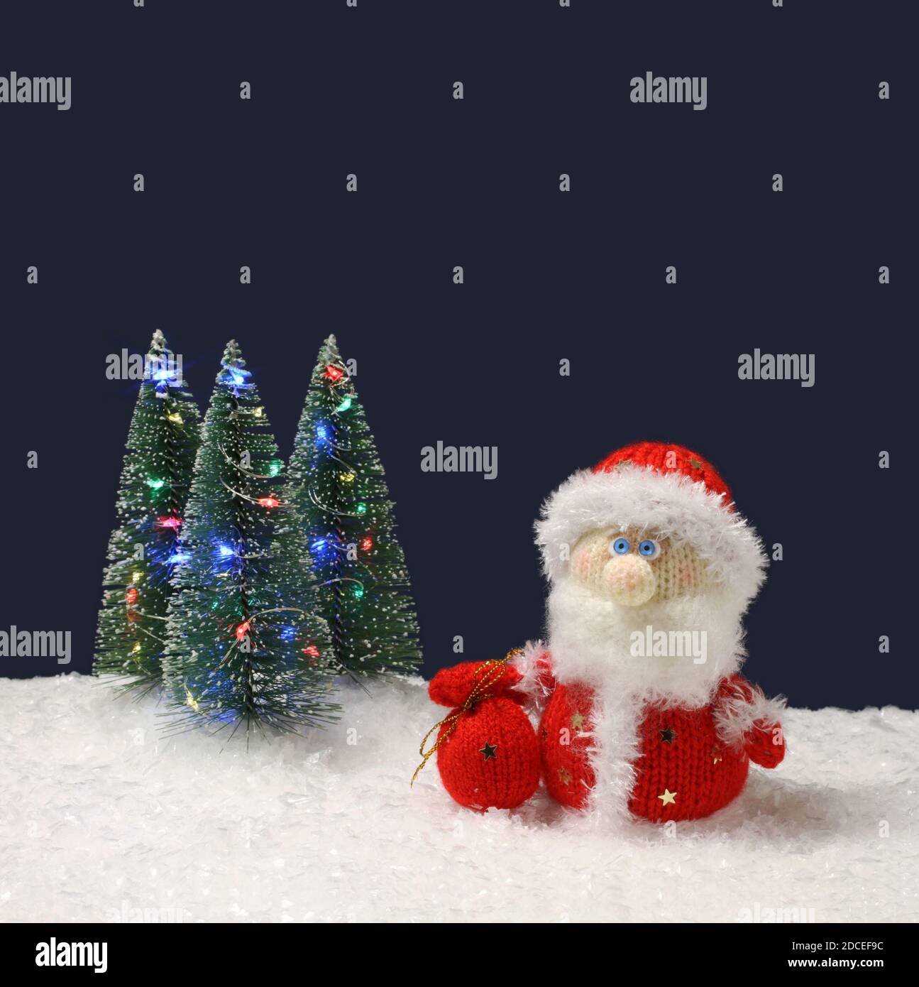 El juguete lana de punto Santa Claus se encuentra junto a los árboles de  Navidad verde juguete con luces sobre la nieve blanca artificial contra  fondo azul marino. Cerrar Fotografía de stock -