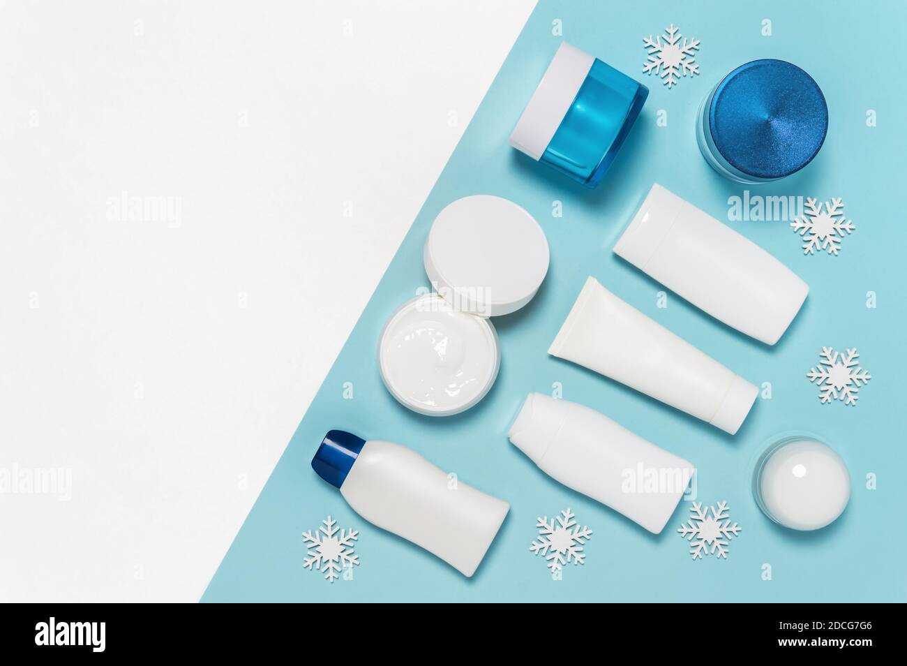 Conjunto de productos cosméticos para el cuidado de la piel de invierno sobre fondo blanco y azul con copos de nieve. Bote de crema, tubo, loción corporal, leche y envase de botella de limpiador Foto de stock