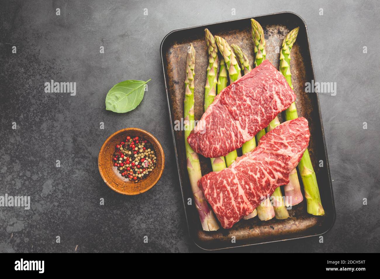Filete crudo con espárragos verdes en bandeja sobre fondo negro, vista superior Foto de stock