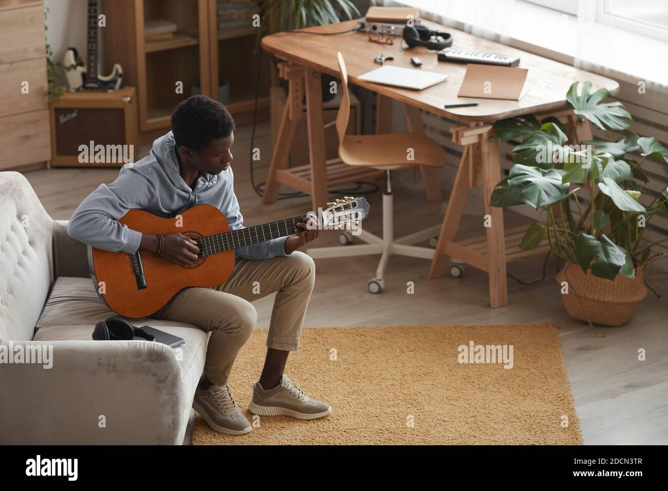 Retrato completo de un joven afroamericano tocando la guitarra acústica mientras se sienta en el sofá en casa, espacio de copiado Foto de stock