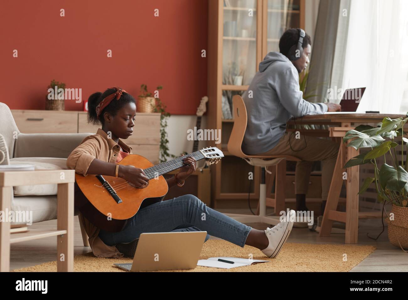 Retrato completo de una músico afroamericana tocando la guitarra y utilizando el portátil mientras se sienta en el suelo en el estudio de grabación, espacio de copia Foto de stock