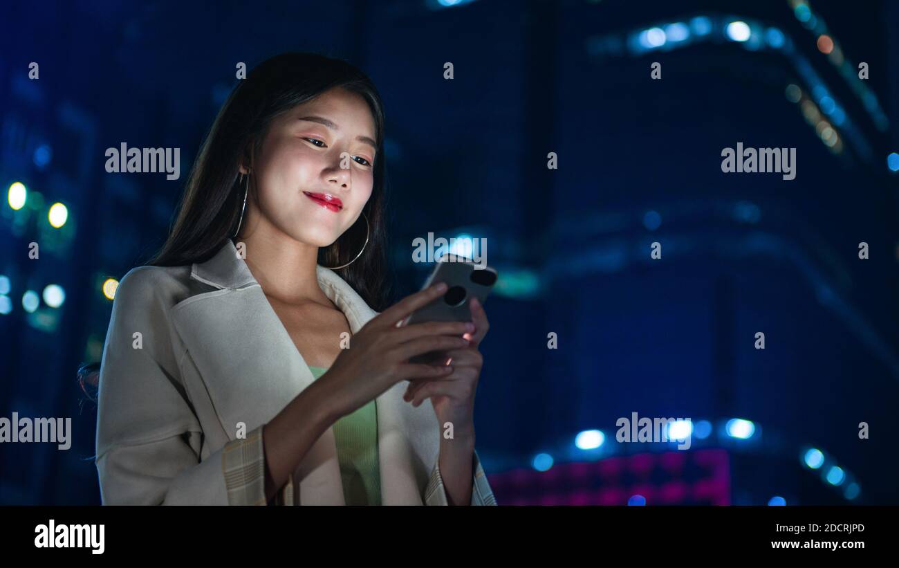 Retrato de una bella mujer asiática sonriente usando un smartphone en la ciudad por la noche Foto de stock
