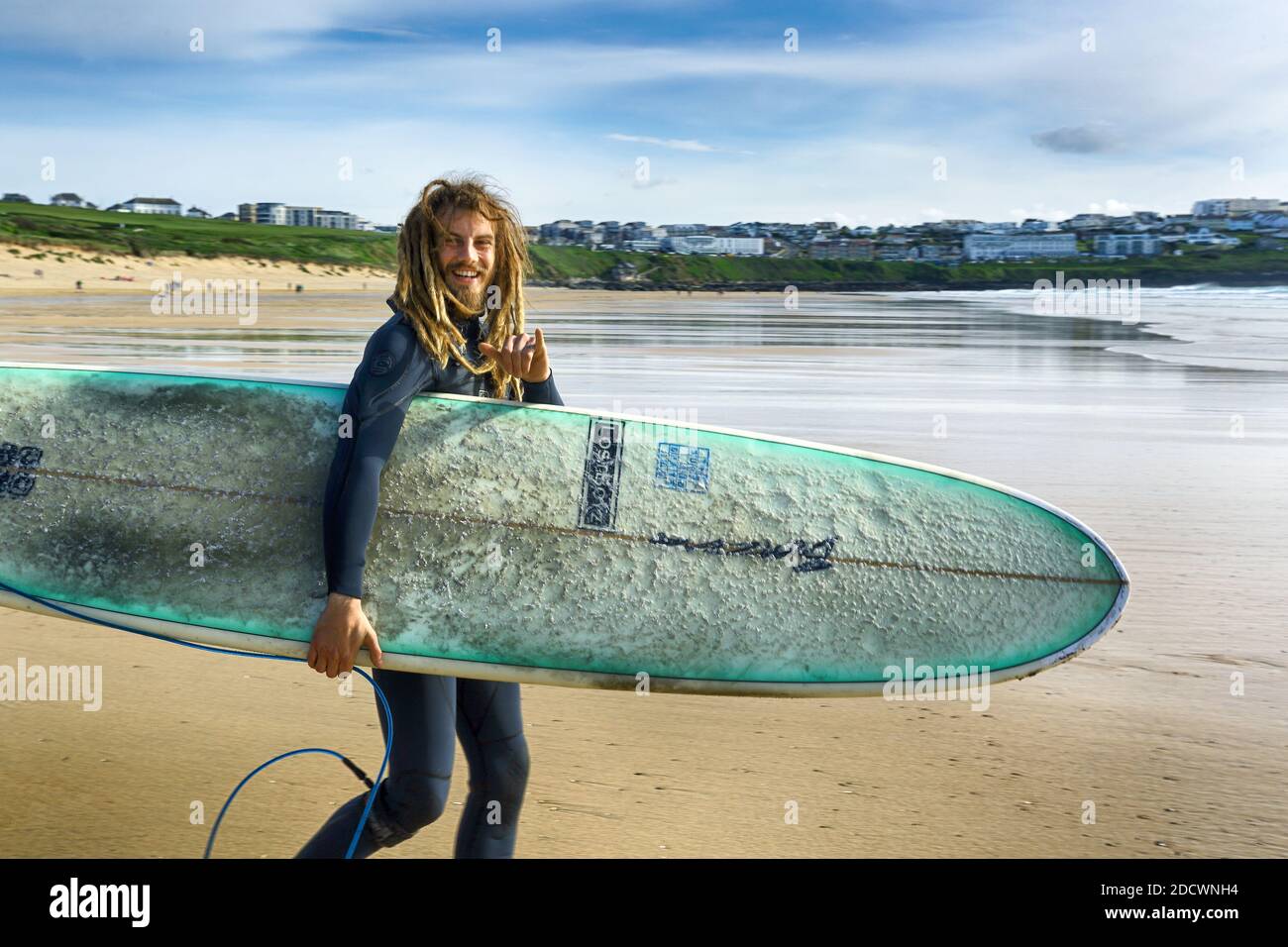 Joven surfista con dreadlocks llevando su tabla de surf y caminando en el mar en la playa de Fistral en Newquay en Cornwall. Foto de stock