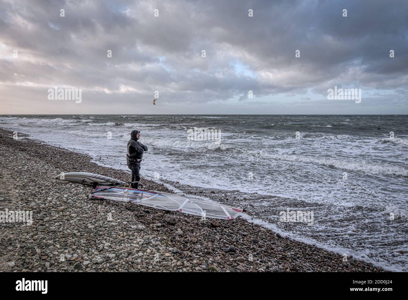 Surfista mayor en traje negro mojado parado en la orilla con su equipo mirando la tormenta de invierno sobre el mar frío, Smidstrup, Dinamarca, Foto de stock