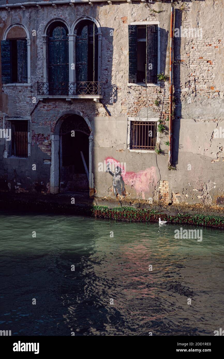 Banksy Street art en Venecia en una casa antigua frente a un canal: La obra de arte retrata a un niño migrante con un chaleco salvavidas y sosteniendo un toque rosa neón. Foto de stock