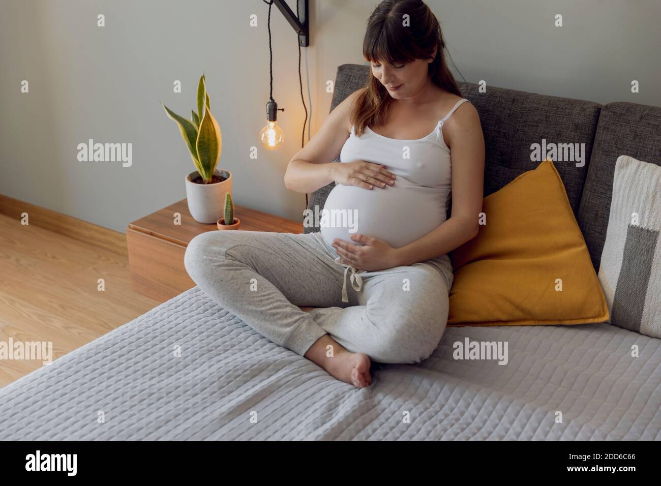 Feliz mujer embarazada sentada en la cama y tocando su vientre en casa Foto de stock