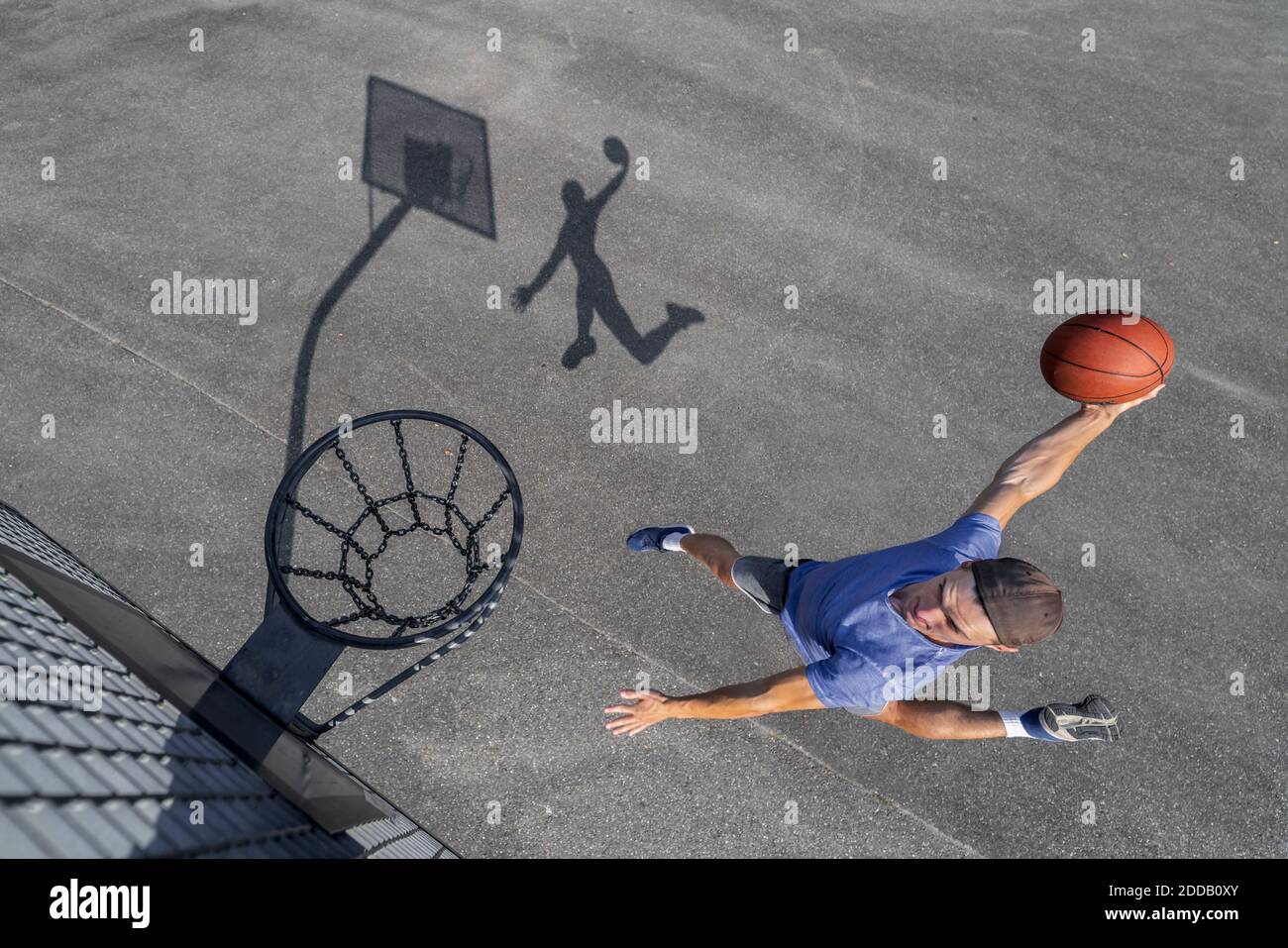 Joven hombre dunking pelota en el aro mientras juega al baloncesto día soleado Foto de stock