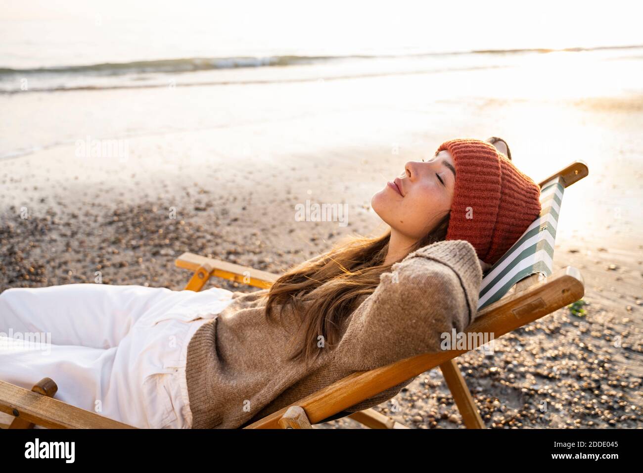 Mujer joven relajándose en silla plegable con los ojos cerrados playa durante la puesta de sol Foto de stock
