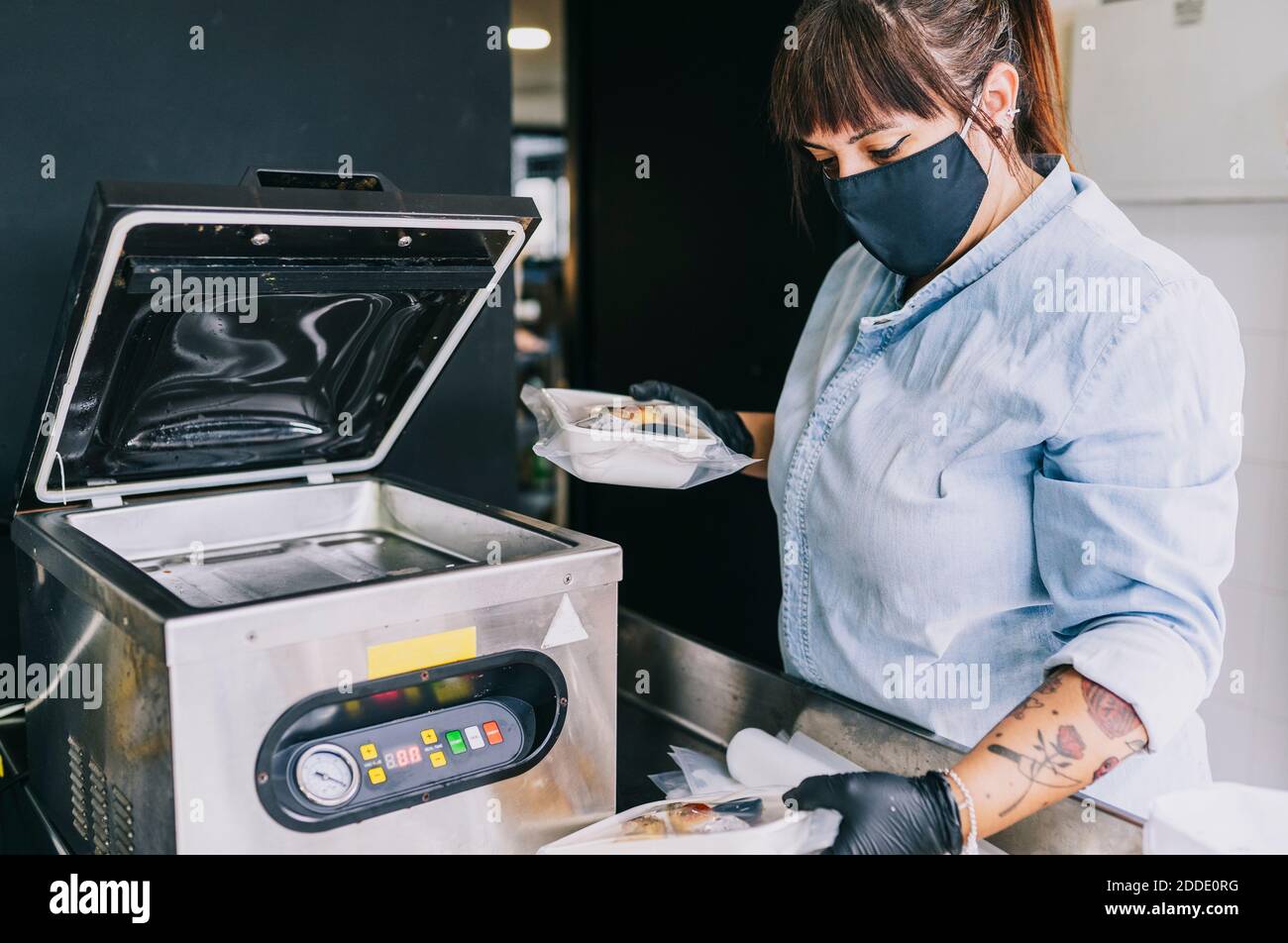 Chef femenino utilizando maquinaria de embalaje hermético mientras se lleva a cabo comida en el restaurante durante la pandemia Foto de stock