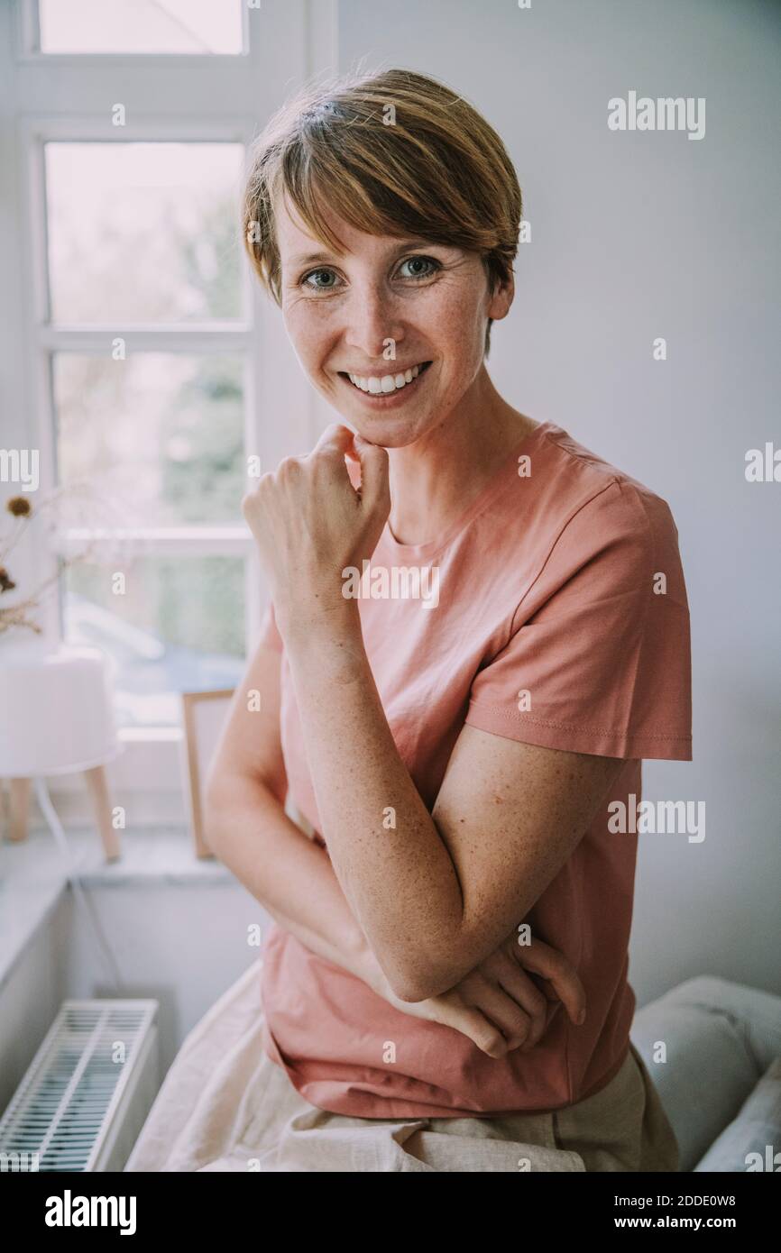 Mujer sonriente con la mano en la barbilla sentada en casa Foto de stock