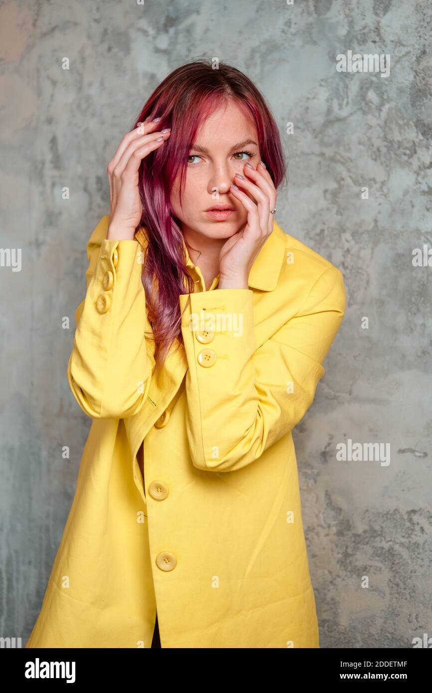 Hermosa mujer con cabello rosa vestido con impermeable amarillo tocando su  cara y posando en el interior cerca de la pared de hormigón Fotografía de  stock - Alamy