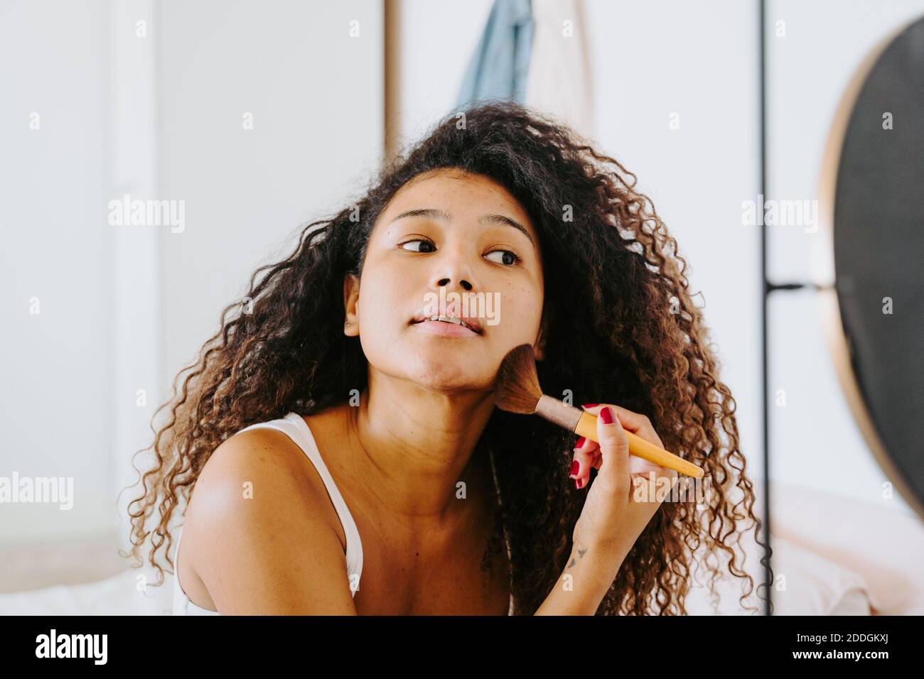 Joven de pelo rizado femenino étnico aplicando rubor de la paleta en mejilla con cepillo cosmético durante el procedimiento diario de maquillaje en casa Foto de stock