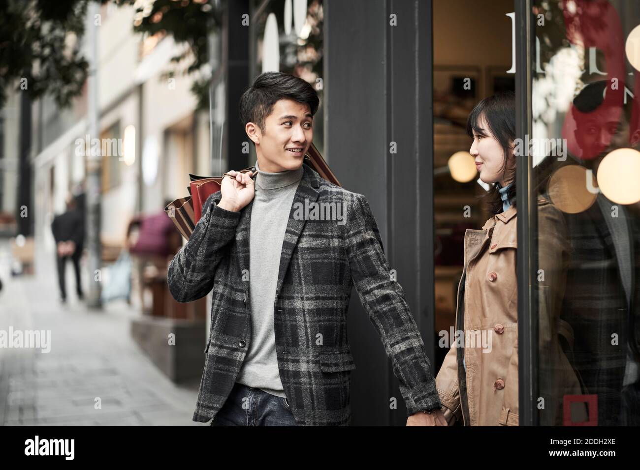 feliz pareja joven asiática de compras en la ciudad caminando fuera de una tienda que lleva bolsas de papel Foto de stock
