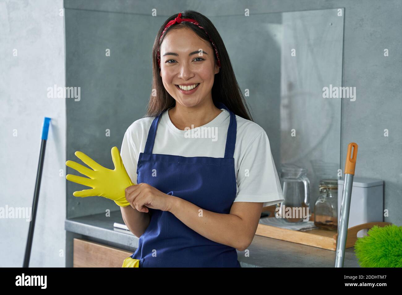 Retrato de una joven alegre, señora de la limpieza sonriendo en la cámara, usando guantes protectores mientras se prepara para limpiar la casa. Las tareas domésticas y el servicio de limpieza, concepto de servicio de limpieza Foto de stock