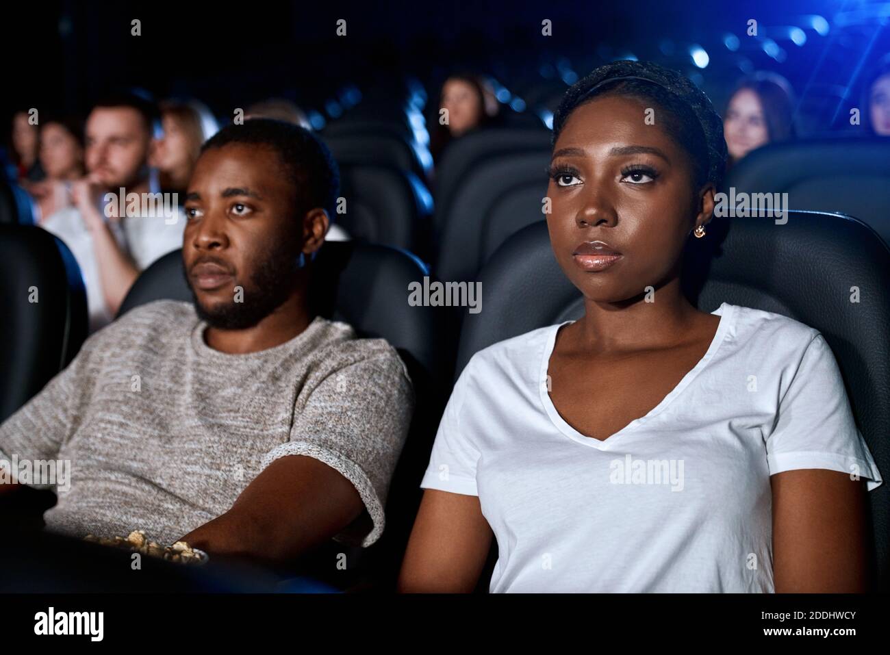 Enfoque selectivo de parejas jóvenes viendo películas en el cine, sentados en cómodos asientos negros, usando camisetas informales. Vista lateral del hombre y la mujer africanos con caras fuertes disfrutando de la Enfoque selectivo de parejas jóvenes viendo películas en el cine, sentados en cómodos asientos negros, usando camisetas informales. Vista lateral del hombre y la mujer africanos con caras fuertes disfrutando de la