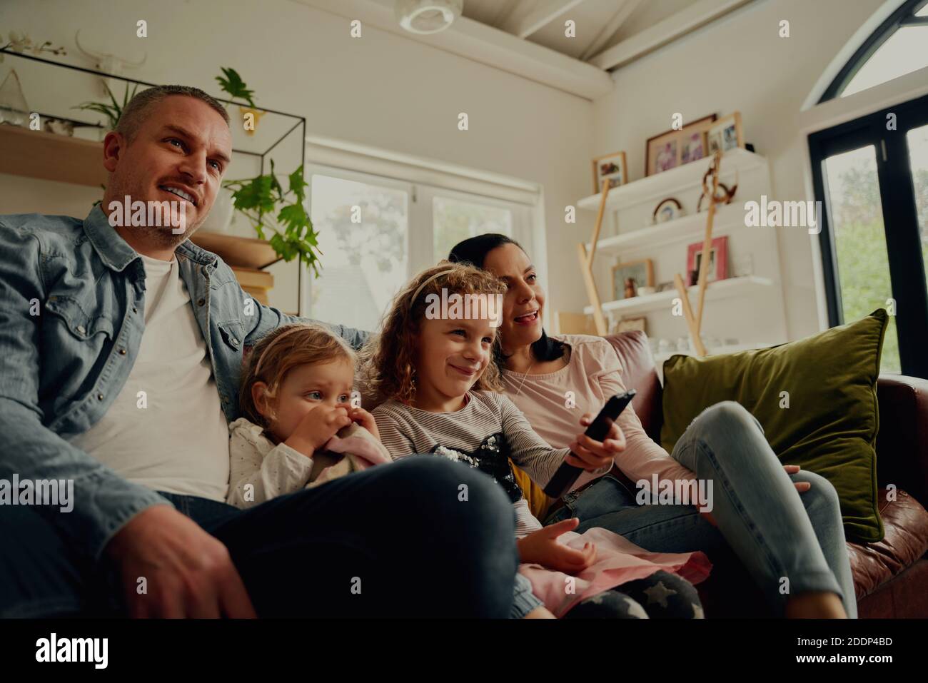 Padres jóvenes sonriendo sentados junto con una hija que controla el mando a distancia en casa Foto de stock