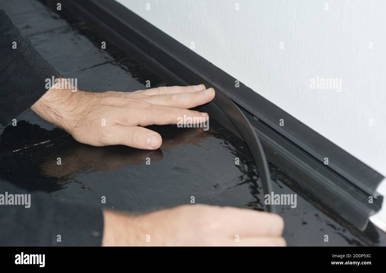 Un hombre está instalando una placa de zócalo de PVC negro a lo largo una  pared de paredes blancas y pisos de madera pintada para ocultar cables  eléctricos detrás de una m