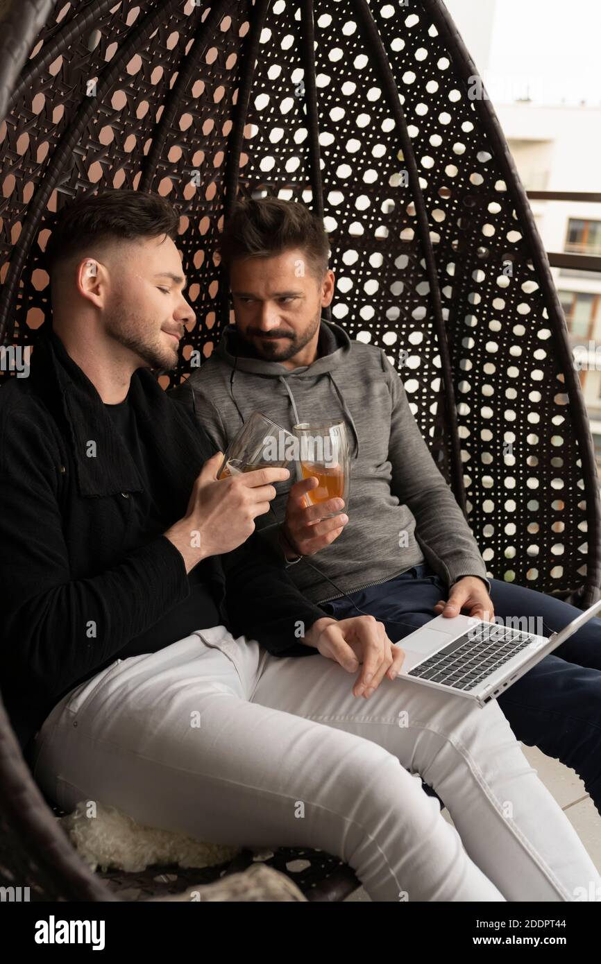 Salud. Joven pareja gay guapo relajarse en el columpio en el balcón con  cerveza y navegar por las redes sociales en el ordenador portátil. Foto de  alta calidad Fotografía de stock -