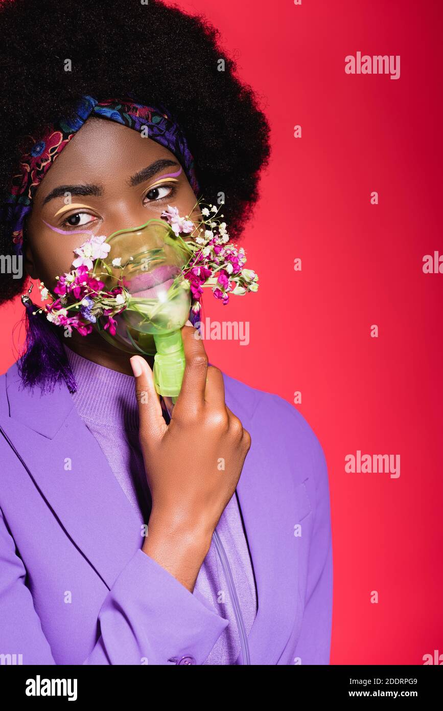 mujer joven afroamericana en traje púrpura con estilo y flores en inhalador aislado en rojo Foto de stock