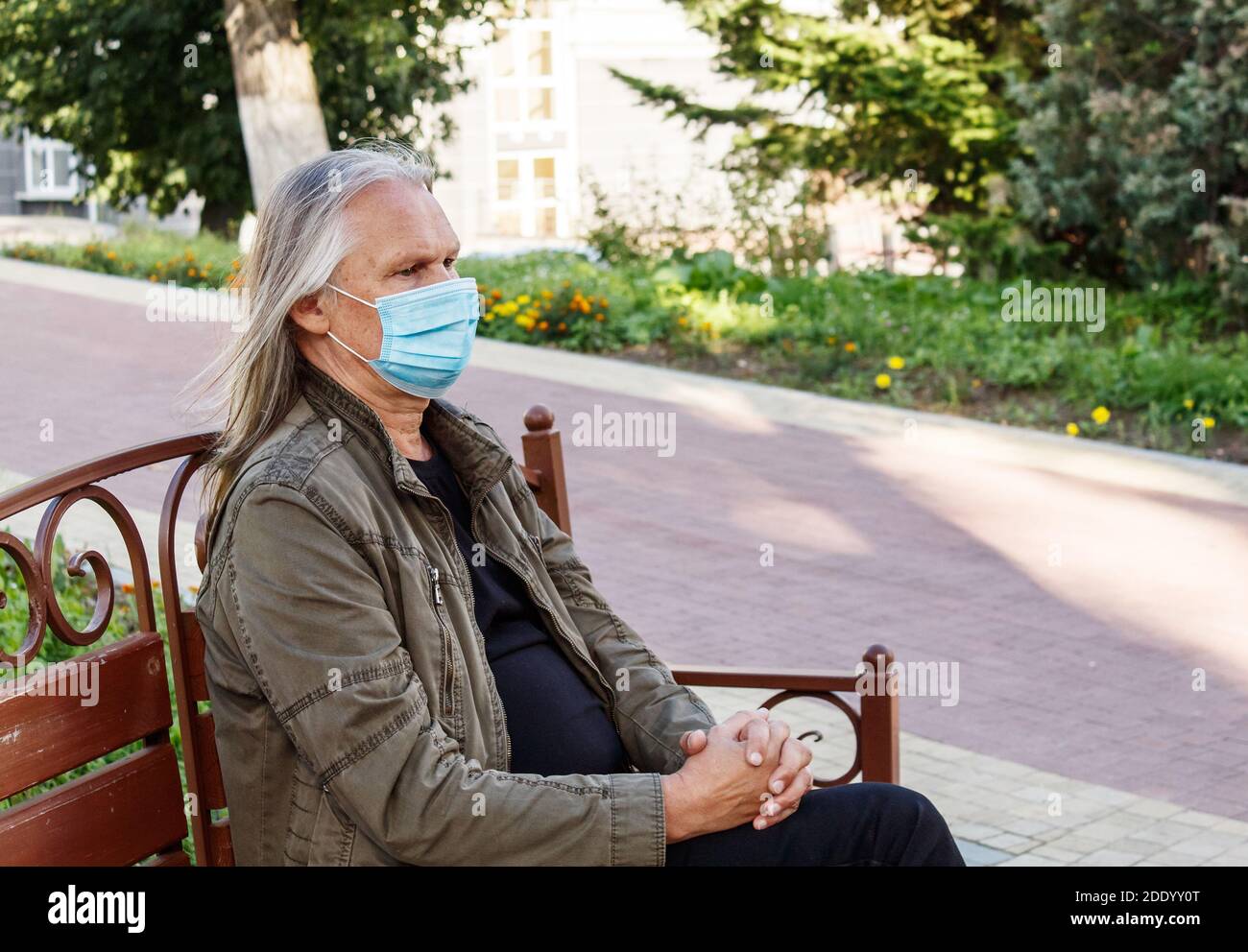 el hombre mayor con mascarilla protectora se sienta en un banco un parque de la ciudad en el soleado día de otoño Foto de stock