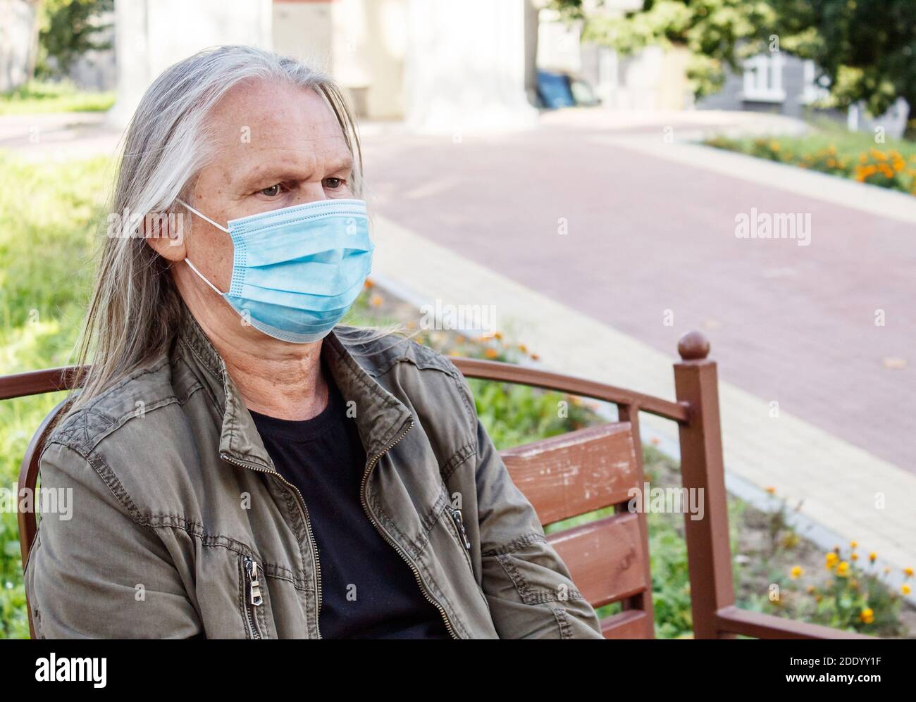 retrato de anciano con pelo largo gris en protector máscara se sienta en un banco en un parque de la ciudad en soleado día de otoño Foto de stock