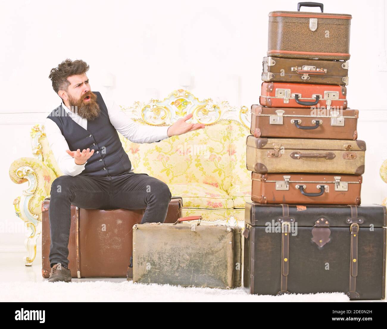 Macho elegante en cara sorprendida se encuentra conmocionado cerca de pila  de maleta vintage. Concepto de equipaje y viaje. Hombre, mayordomo con  barba y bigote entrega el equipaje, fondo interior blanco de