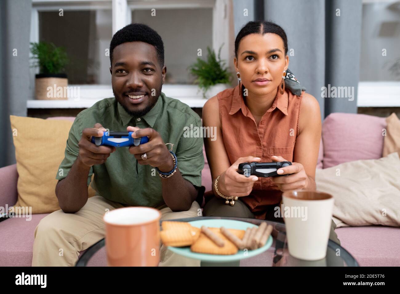 Feliz adolescente de etnia africana y chica de raza mixta sentada en el sofá contra la ventana en la sala de estar y jugar a videojuegos Foto de stock