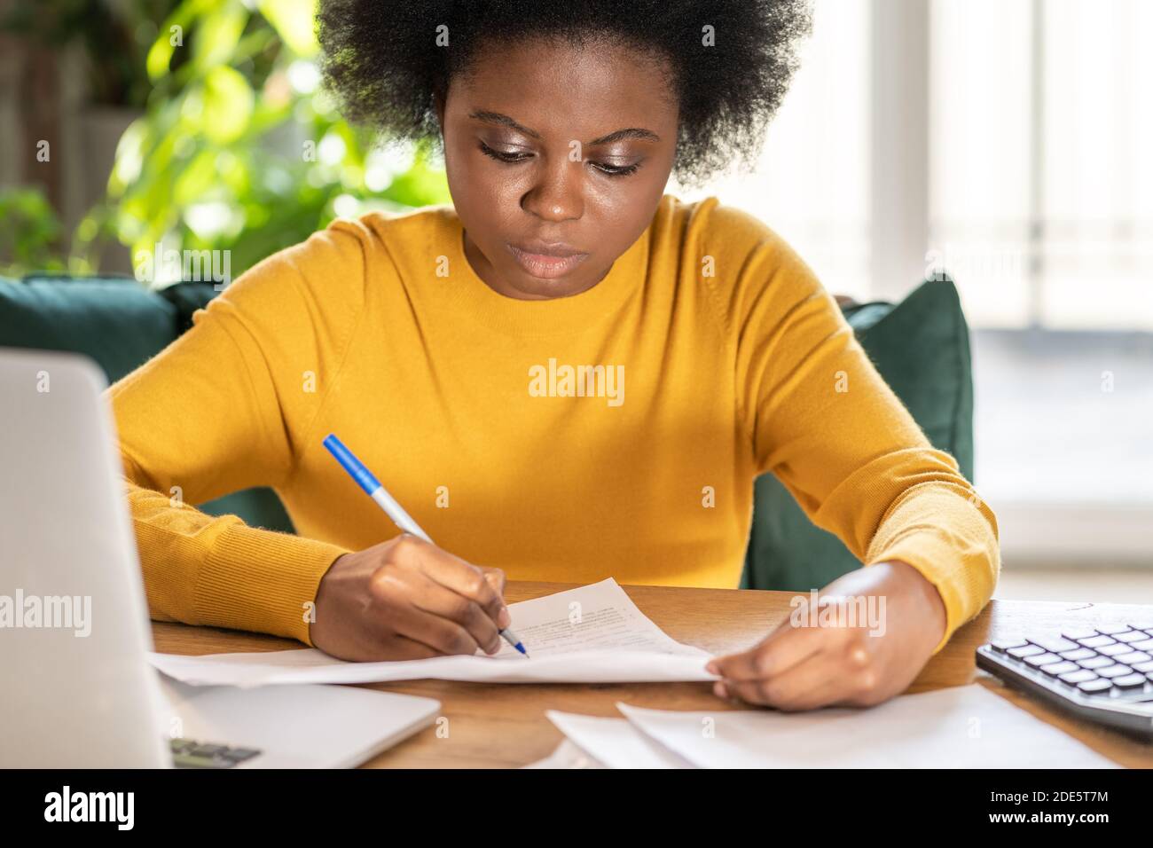Mujer negra empleada o gerente de trabajo remoto en casa durante el cierre, toma notas con el lápiz, trabaja con documentos en papel. Trabajador de oficina durante una pande Foto de stock