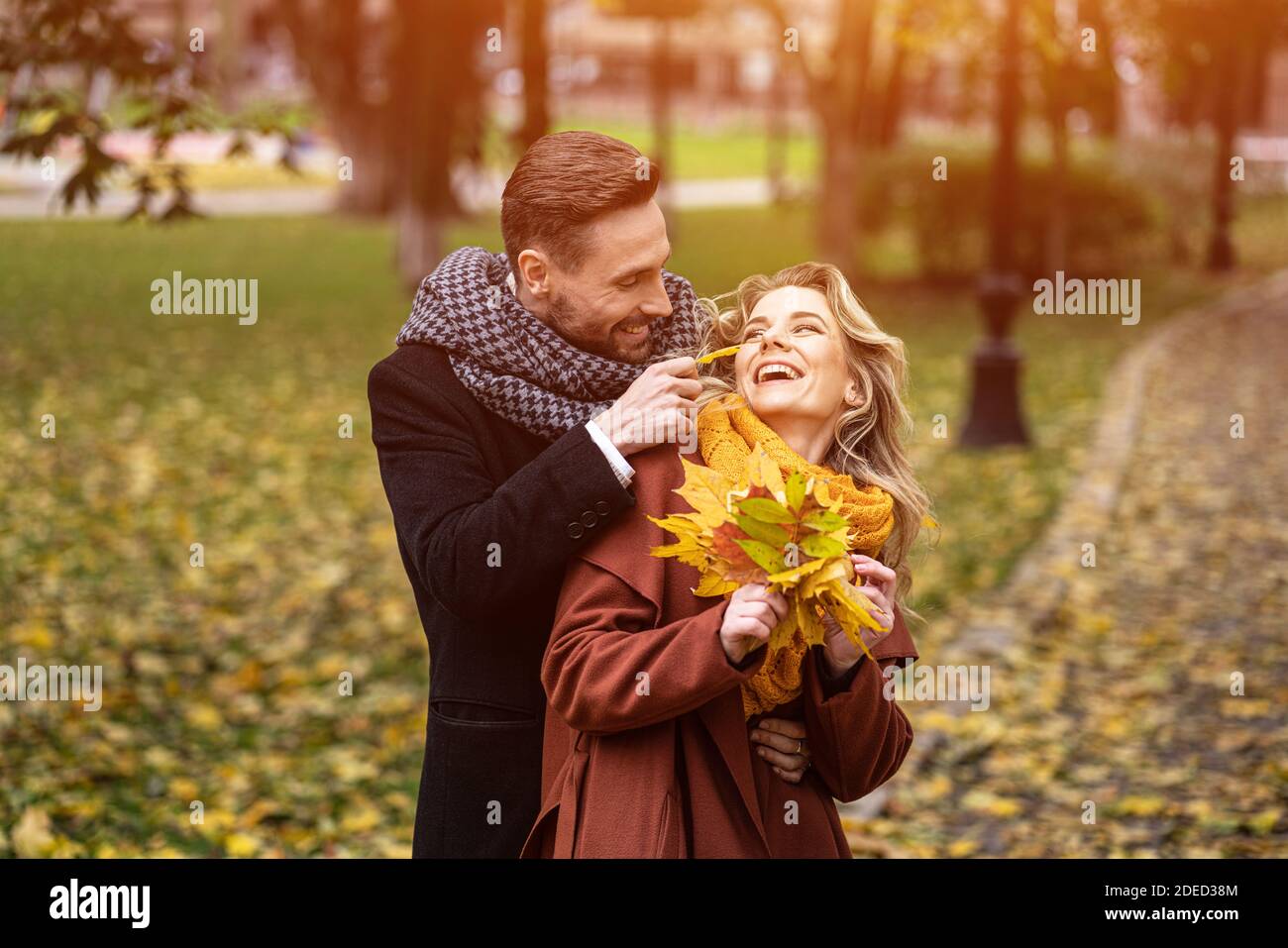 Feliz gente joven enamorada, hombre abrazando a una mujer desde atrás acariciando su mejilla, feliz pareja caminando en un parque de otoño con elegantes abrigos y escogiendo Foto de stock
