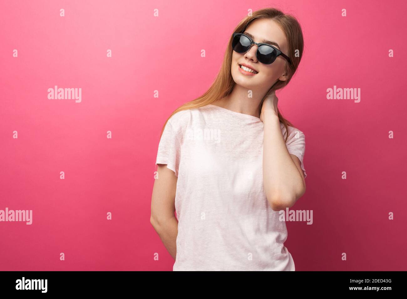 Hermosa joven rubia chica en gafas de pie sobre fondo rosa con jeans, rosa superior sonriente nieve sonrisa blanca, con gafas negras y mirar Foto de stock