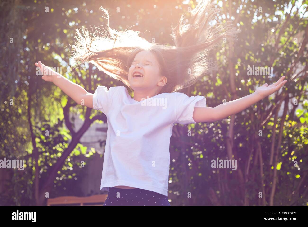 Feliz chica joven sola, pequeño niño alegre en edad escolar saltando, volando pelo disipado, brazos extendidos, luz solar, retrato al aire libre. La infancia Foto de stock